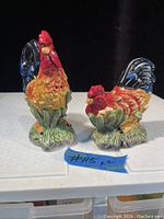 Front angle view of ceramic rooster and hen salt and pepper shakers on white surface with black background.