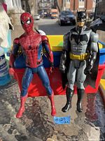 Photo shows Spider-Man and Batman action figures standing side by side outdoors. Spider-Man is red and blue, Batman is gray and black with a yellow belt.
