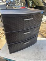 Front angle view of the black Sterilite 3-drawer dresser on a concrete surface with cars in background, showing the woven texture drawer fronts and silver handles.