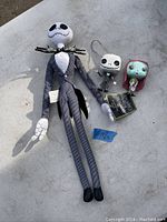 Photo showing one large Jack Skellington stuffed doll with pinstriped suit and bat bow tie, and two smaller figurines of Jack Skellington and Sally characters in good condition.