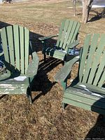 Three green wooden Adirondack chairs placed outdoors on grass with noticeable paint wear and damage to wood.