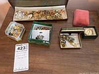 Full view of the assortment showing jewelry in open boxes and trays on a wooden surface. Includes assorted pins, brooches, and chains.