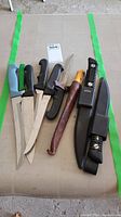 Overview of 7 knives including various styles and handles, laid on surface with green tape edges.