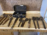Overview of assorted hammers and pipe wrenches laid out on wooden countertop with black metal toolbox containing chainsaw tools.