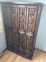 Front and corner view of vintage highboy dresser showing detailed carved floral and scroll designs on wood panel doors and drawers.
