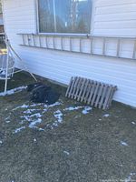 Aluminum ladder resting outdoors against a white building wall with other miscellaneous items nearby including a wooden gate or fence panel.