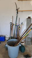 Full view showing various garden tools including rakes, shovels, some hand tools, watering can, and a trash can inside a garage.