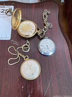 Three pocket watches on a wooden surface: gold-tone Caravelle with chain, gold-tone English Time Company hunter case watch with ornate chain, silver-tone Santima watch showing wear.