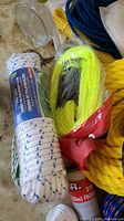 Photo showing coiled white polypropylene rope with blue tracers, coiled yellow rope with metal hook, and other assorted ropes on a light surface.