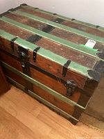 Overall angled view of the wooden steamer trunk showing the green slats and brown wood panels with black metal reinforcements and hardware.