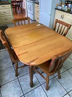 Wooden dining table with six matching chairs around it in a kitchen setting. The table has rounded corners and is medium brown in color.