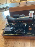 Side angle of the vintage Singer sewing machine showing the black body, gold detailing, spool of white thread on top, foot pedal wrapped in cord, and wooden base. The carrying case is visible in the background.