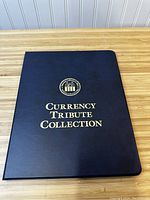 Closed black collector's folder with gold embossed lettering 'Currency Tribute Collection' on bamboo surface.
