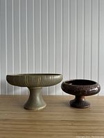 Pair of pedestal planters or vases with mid-century modern design, one olive green matte textured, one dark brown glossy, on a wooden surface
