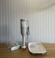 White Braun handheld electric mixer with power cord wrapped and beaters in a plastic bag, placed on a wood surface with a white tray beside them.