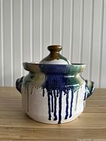Front view of vintage ceramic pot with lid, showing dripping paint glaze in blue, green, and brown over white, with two side handles