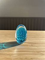 Front view of the blue Murano glass egg paperweight showing bubble design inside, placed on wooden surface with natural sunlight casting a shadow.