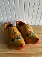 Pair of yellow toned wooden clogs with hand painted Dutch scenes including a windmill, houses, and a boat, tipped and edged in red paint