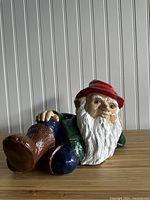 Side angle of the vintage porcelain gnome figurine lying down showing the detailed face, white beard, red hat, green shirt, brown boots and textured base.