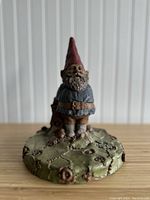 Front angled view of the forest gnome figurine on its base showing facial features, red pointed hat, blue shirt, brown belt, beige pants and textured leaf and acorn base.