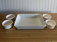 White ceramic rectangular baking dish centered with four round ramekins arranged around it on wooden surface, vertical bead board background.