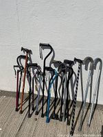 Photo showing 17 assorted walking stick canes standing upright against a wall outdoors, varied colors and handle styles visible.