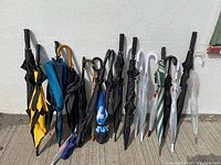 Full view of 13 assorted umbrellas showing a mix of black, blue, yellow, green, and clear colors, with some curved and straight handles leaning against a white wall.