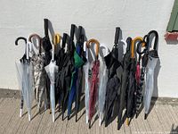 Photo showing 22 assorted umbrellas arranged upright against a wall, displaying various handle types and colors including black, white, clear, and patterns.