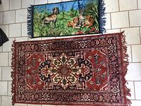 Two assorted carpet tapestry rugs laid on tiled floor. One with animal scene and blue fringe, the other with traditional red geometric pattern and fringe.