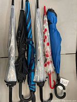 Full view of 6 assorted umbrellas standing upright wrapped in plastic or folded, showing different colors and handle styles.