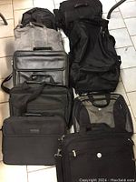 Nine assorted bags displayed on floor, including laptop bags and duffle bags in black, gray, and olive colors, some with shoulder straps and handles.