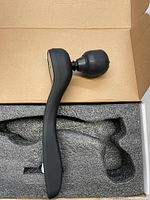 Black handheld massage gun inside cardboard box with foam padding for protection, showing ergonomic handle and rounded massage head