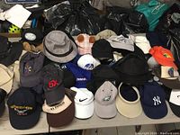 Full view of assorted hats laid out on table: baseball caps featuring various logos, fedoras, and sun hats in multiple colors and styles such as black, beige, orange, and grey.