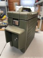 Photo showing angled side front view of the green military dispenser container with handle and latch closure, highlighting the rugged design and spout area.