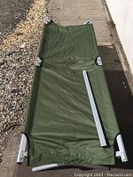 Full view of folded-out green fabric camping cot with aluminum frame lying flat on ground showing fabric and frame ends.
