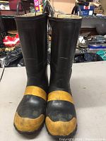 Front view of the pair of black firefighter boots with yellow toe caps and strap details, standing upright with pull handles visible.