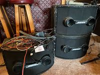 Three Bose speaker modules (stack of two on right, one on left) with cables coiled on top, paired with speaker stand base and microphones on floor