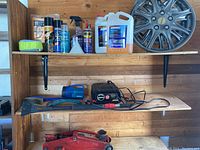 Shelf containing cleaning supplies, Schumacher battery charger, three hubcaps on upper shelves, Mastercraft hydraulic jack on bottom shelf