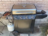Front view of Char-Broil grill with stainless steel hood, control panel with four knobs, side burner, grilling utensils on left shelf, propane tank