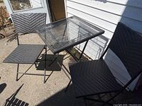 Two black woven folding chairs with a matching rectangular side table with tempered glass top on outdoor concrete surface.