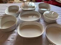 Lot of various CorningWare French White stoneware baking dishes and casseroles with clear glass lids on a table.