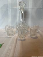Front view of cut glass decanter with stopper surrounded by five matching shot glasses on a light textured cloth.