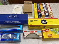 Various boxes of aluminum foil, sandwich bags, snack bags, parchment paper, and other kitchen wraps packed together on a table.