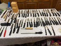 Wide view showing extensive assortment of knives laid out on table with a wooden knife block and black knife sharpener.