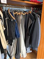 Photo of multiple men’s suit jackets and pants hanging in a closet on hangers, shown in various dark and neutral tones including black, brown, and blue.