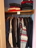 Wide shot of closet showing hanging men's sweaters in large size and folded sweaters on shelf above.