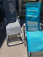 Two foldable outdoor loungers, grey and blue, shown side by side on pavement with wooden deck background.