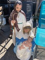 Full view of all three resin religious figurines placed outside on pavement near chairs, showing Joseph standing behind Mary holding baby Jesus.