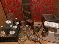 View of vintage meat grinders, Moulinex grinder, wine jugs, canning jars, and wooden slicer against red patterned wall.