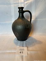 Front view of black clay vessel with handle and round body on white textured background.